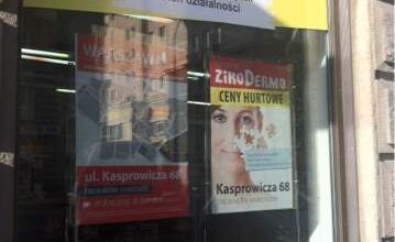 Widok zewnętrzny apteki z plakatami informacyjnymi i reklamowymi, w tym dla apteki internetowej e-zikoapteka.pl, w centrum miasta, z widoczną ulicą Kasprowicza 68.