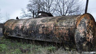 Zardzewiały stary zbiornik na paliwo na zaniedbanej powierzchni, porośnięty trawą i kasztanami, w tle drzewostan, symbol przemijania i zaniedbania w krajobrazie przemysłowym.
