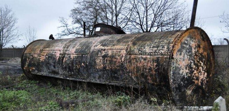 Zardzewiały stary zbiornik na paliwo na zaniedbanej powierzchni, porośnięty trawą i kasztanami, w tle drzewostan, symbol przemijania i zaniedbania w krajobrazie przemysłowym.