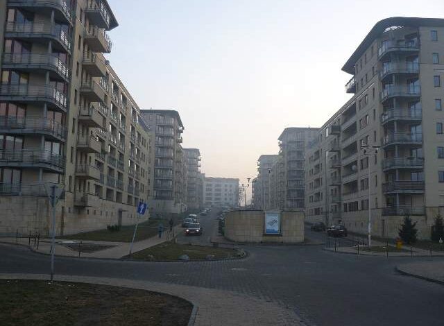 Nowe osiedle mieszkaniowe w mieście, budynki wielopiętrowe z balkonami i ulicą w centrum, panorama nowoczesnej architektury, codzienne życie mieszkańców, komfort i funkcjonalność.