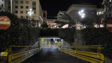 Podziemny parking podziemny w centrum miasta, oświetlony nocą z widokiem na budynki i rzeźbę smoka, otoczony zielenią i oznakowanymi słupkami z barierkami.