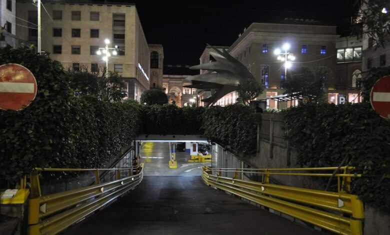 Podziemny parking podziemny w centrum miasta, oświetlony nocą z widokiem na budynki i rzeźbę smoka, otoczony zielenią i oznakowanymi słupkami z barierkami.