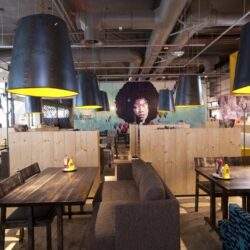 Lampy przemysłowe nad stolikami w nowoczesnej restauracji, wnętrze z muralem kobiety Afro na ścianie, przytulna atmosfera, drewniane elementy, optymalny klimat do spotkań i relaksu, optima optymalizacja dla restauracji.