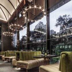 Eleganckie wnętrze restauracji z nowoczesnym oświetleniem, przytulnymi zielonymi kanapami i dużymi oknami z widokiem na zieleń, idealne na spotkania i relaks.