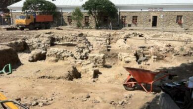 Prace archeologiczne na budowie z ceglaną chatą w tle, przygotowania do prac budowlanych, widok na wykop pod fundamenty, narzędzia budowlane i maszyny na placu budowy, zarys starego domu.
