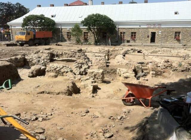 Prace archeologiczne na budowie z ceglaną chatą w tle, przygotowania do prac budowlanych, widok na wykop pod fundamenty, narzędzia budowlane i maszyny na placu budowy, zarys starego domu.