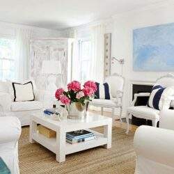 Przytulny salon w jasnych kolorach z eleganckimi białymi meblami, dekoracyjnymi poduszkami i kwiatami na stole, idealny do relaksu i spotkań towarzyskich.