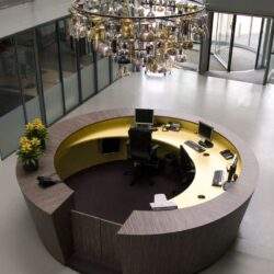 Nowoczesny recepcjonistyczny stojak z komputerami i telefonami w minimalistycznej, eleganckiej przestrzeni biurowej ze szklanymi ścianami i dużym żyrandolem z dekoracyjnymi elementami metalowymi.