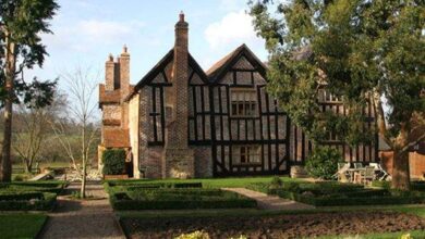 Stary dom w stylu angielskim w ogrodzie z kwietnikami i dużymi drzewami, piękny widok na zielony teren i częściowo zachmurne niebo.