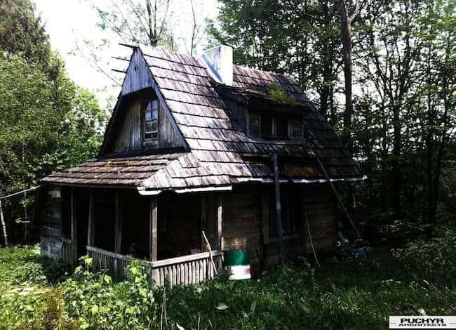 Stara, zaniedbana chałupa drewniana w otoczeniu lasu, niska kondycja budynku, z wrakami i zaroślami wokół, ukazująca stare budownictwo wiejskie, idealne dla miłośników historii i architektury.