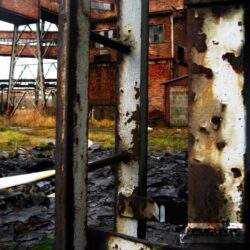 Zardzewiała metalowa bariera na tle starej opuszczonej fabryki przemysłowej, zniszczony i zbutwiałe elementy strukturalne, postindustrialny krajobraz, symbol zaniedbania i przemijania, kluczowe dla industrialnej estetyki, opuszczone miejsce, depresja i nostalgiczne wspomnienia, optymalne rozwiązania dla odnowy terenów przemysłowych, PaniDyrektor.pl.