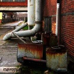 Brudne, zardzewiałe urządzenia przemysłowe i rury na tle ceglanej ściany, w scenerii miejskiej, sugerujące zaniedbanie, przemysłowy klimat i potencjalny brak optymalizacji przestrzeni miejskiej.