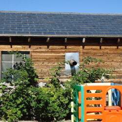 Stara drewniana chatka z panelami słonecznymi na dachu, otoczona zielenią, w otwartym ogrodzie z placem zabaw, idealna dla osób poszukujących ekologicznych rozwiązań mieszkaniowych.