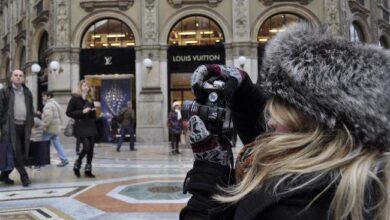 Kobieta robi zdjęcie w centrum handlowym, zimowa czapka i rękawice, elegancki shopping street z markami luksusowymi w tle.