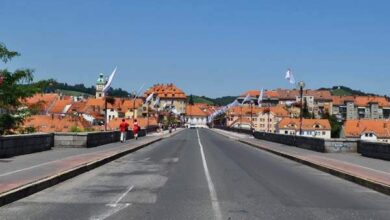 Bridge nad rzeką w mieście z zabytkową architekturą i czerwoną dachówką, w słoneczny dzień, obok spacerujący ludzie, widok na wzgórza w tle, idealne miejsce na zdjęcie dla portalu PaniDyrektor.pl.
