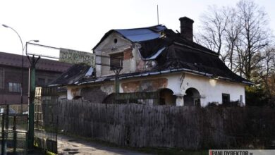 Zrujnowany budynek mieszkalny z uszkodzonym dachem i konstrukcją, wymaga renowacji i odbudowy, na tle drzew i innych budynków w okolicy.