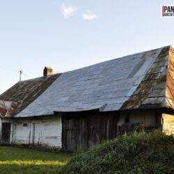 Stary wiejski dom z metalowym dachem, drewnianymi ścianami i ogrodem, na tle nieba w słoneczny dzień, element architektury tradycyjnej i wiejskiej, idealny do projektów rewitalizacji i architektury krajobrazu.