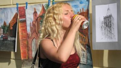 Obraz młodej kobiety pijącej napój podczas wystawy sztuki na zewnątrz, w tle zawieszona sztuka i rysunki architektoniczne.