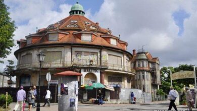 Willa z czerwonym dachem i oknami w stylu historycznym, otoczona drzewami, w centrum miasta, na tle nieba z chmurami, reprezentująca architekturę z początków XX wieku.