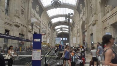 Przestrzeń dworca kolejowego w wysokim, zabytkowym stylu z ludźmi oczekującymi na pociąg i podróżującymi z bagażami, ukazująca architekturę i atmosferę podróży.