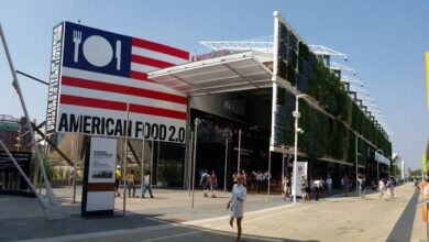Nowy budynek targowy z logo American Food 2.0 na zewnątrz, nowoczesna architektura, duże przeszklenia, zielone elewacje, tłum zwiedzających, idealne miejsce na wydarzenia branżowe, design i funkcjonalność.