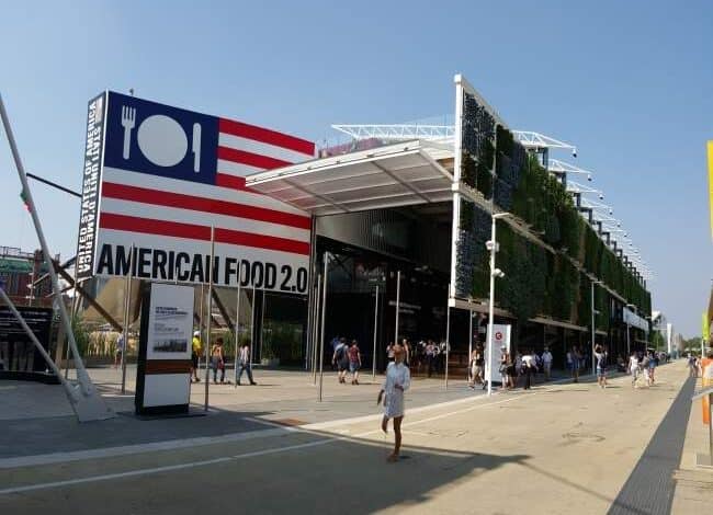 Nowy budynek targowy z logo American Food 2.0 na zewnątrz, nowoczesna architektura, duże przeszklenia, zielone elewacje, tłum zwiedzających, idealne miejsce na wydarzenia branżowe, design i funkcjonalność.