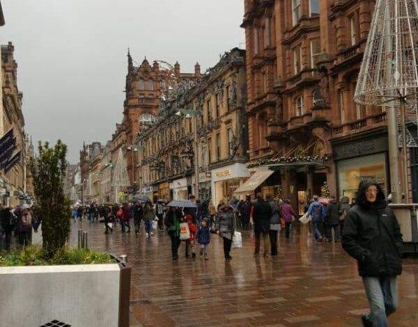 Londyńska ulica w deszczowy dzień z tłumem przechodniów, stare kamienice i miejskie dekoracje, atmosfera miejskiej burzy, mokre chodniki, popularny klimat miasta, świąteczne ozdoby na ulicach, dynamiczny krajobraz urbanistyczny, stylowa architektura.