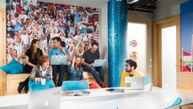 Odpoczywająca grupa młodych ludzi w nowoczesnym coworkingu z kolorową ścianą pełną zdjęć, pracują na laptopach, atmosfera sprzyja kreatywności i współpracy w dynamicznym środowisku pracy.