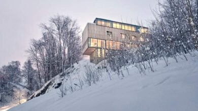 Nowoczesny dom na zboczu zimowego lasu, widok z dołu, duże okna, minimalistyczny styl, zimowa sceneria, ostre krawędzie budynku, luksusowa architektura, panorama, nastrojowe światło, nowoczesne rozwiązania architektoniczne.