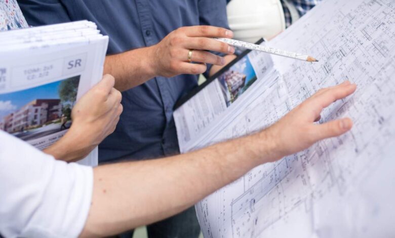 Prezes i architekt omawiający projekty architektoniczne na placu budowy, pojawia się dokumentacja budowlana, plany mieszkań i wizualizacje. Współpraca architektoniczna i nadzór inwestycyjny.
