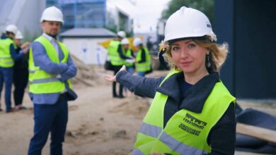 1. kobieta na budowie w kamizelce odblaskowej i kasku, wskazuje na plac budowy, grupa pracowników w tle, architektura, realizacja projektu, kontrole budowlane, bezpieczeństwo, optymalizacja procesu.