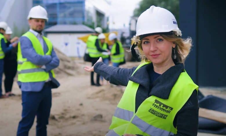 1. kobieta na budowie w kamizelce odblaskowej i kasku, wskazuje na plac budowy, grupa pracowników w tle, architektura, realizacja projektu, kontrole budowlane, bezpieczeństwo, optymalizacja procesu.